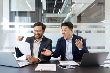 Two business colleagues behind paperwork rejoicing and celebrating victory triumph and successful achievement results. Men in business suits sitting at desk reviewing financial investment reports.