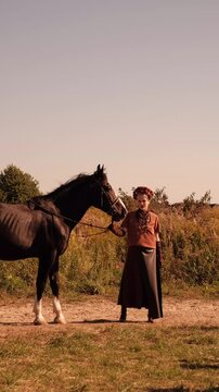 A serene video of a woman in traditional ethnic clothing standing and holding a brown horse by the reins, with an open field stretching behind them.