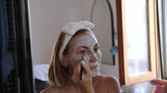 beauty skincare woman applies a cleansing clay mask to her face near the mirror facial skin care home spa