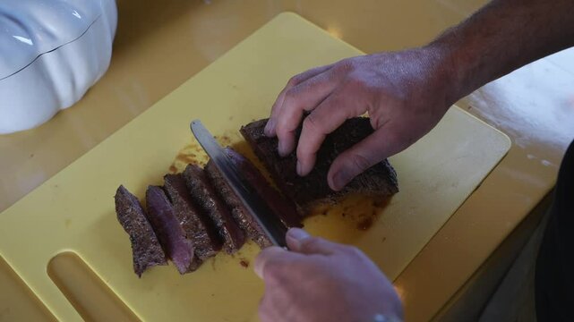 A man is cutting a piece of meat on a yellow cutting board. The meat is sliced into thin pieces