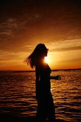Fototapeta premium silhouette of a young girl in sunglasses relaxing by the sea during sunset. The wind blows the long hair of a girl at sunset by the sea