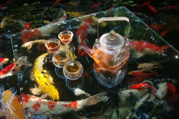 Relaxing tea set on glass table by serene pond filled with colorful koi fish during a sunny day