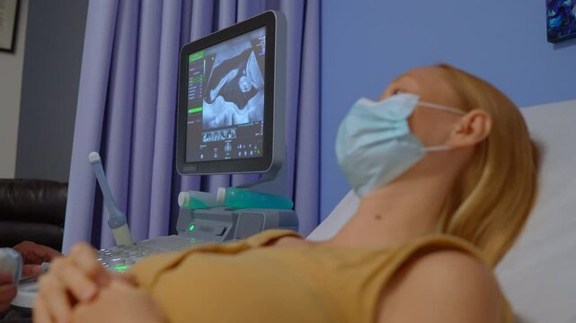 Pregnant woman wearing a medical mask in her late stage of pregnancy is undergoing an ultrasound check at the doctor's office. Expecting mother receiving prenatal care while prioritizing health and