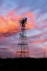 a silhouette of a windmill against a vibrant sunset sky. The sky is painted with shades of orange, red, and yellow, creating a striking contrast with the dark outline of the windmill.