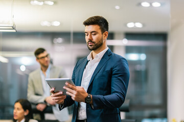 Portrait of young handsome businessman standing in his office in front of his colleagues with tablet in his hands.