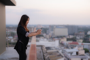 Businesswoman is using her smartphone while leaning on the railing of a rooftop, overlooking a...