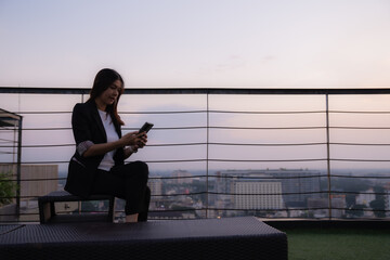 Businesswoman sitting on a rooftop at dusk, using her smartphone while enjoying a breathtaking city skyline bathed in evening light for work and reflection