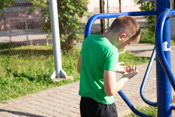 Obraz premium A teenage boy in a green shirt pauses his outdoor workout, checking his hands near a blue exercise machine. The setting is a sunny park with paved paths.
