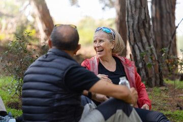 Fototapeta premium An elderly woman laughs with friends at a relaxing outdoor gathering in a serene forest, sharing stories and enjoying a peaceful break after a trekking hike in the woods. Horizontal.