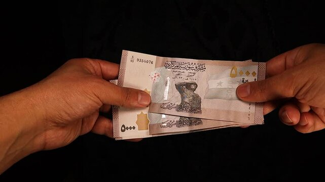 Hand holding a bundle of 5000 Syrian pound banknotes passes it to another hand in a seamless transaction. Represents Syrian currency exchange, financial transfer, business dealings in a clean setting.
