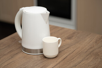 Water is poured from a white electric kettle into a white cup on a wooden table in a cozy kitchen environment