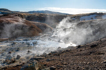 Geyser steam is coming out of the ground in the Seltun Geothermal Area. The ground is covered in rocks.