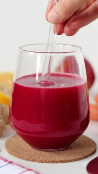 Hand is mixing red beetroot juice placed on table with carrots, ginger, and lemon. Vertical video. Close-up.