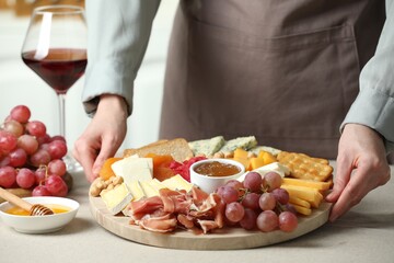 Woman holding board with different types of cut cheese and other snacks at light textured table, closeup