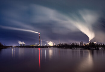 Fototapeta premium Industrial Plant Emitting Smoke Reflected in Calm Water During Twilight. A nighttime industrial scene with vivid reflections on water and atmospheric smoke flows. Orlen Lietuva in Lithuania