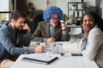 Young African American female employee covering mouth with hand while laughing at strict clown boss...