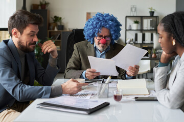 Mature male director of business company wearing clown wig looking through financial paper...