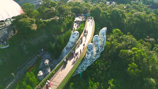 The Golden Bridge in Ba Na Hills, Vietnam, features massive hands supporting the walkway, surrounded by lush greenery and attracting numerous visitors. Aerial drone view.
