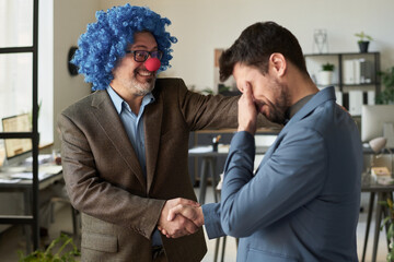 Smiling mature male clown boss keeping hand on shoulder of young subordinate and handshaking with...
