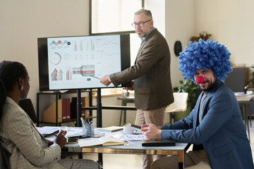 Young smiling businessman in clown wig looking at camera while sitting by workplace against mature...