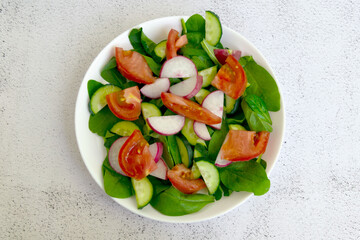 Raw fresh green healthy organic salad of spinach, cucumber, tomato and radish in white bowl on table top view on grey background.