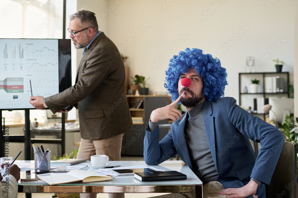 Poster young pensive businessman in clown wig sitting by desk in front of speaker explaining data on intera