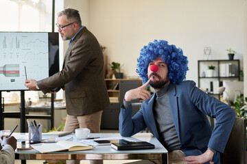 Young pensive businessman in clown wig sitting by desk in front of speaker explaining data on...
