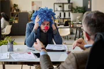 Young stressed businessman in clown wig and nose sitting in front of boss and holding head in hands...