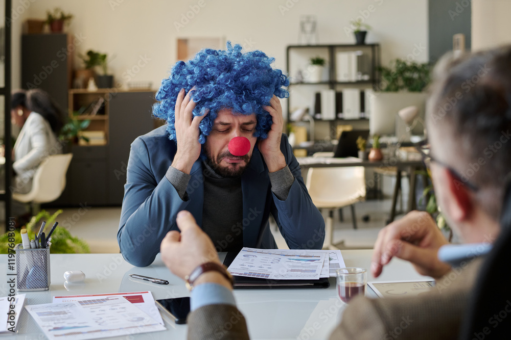 Aufkleber upset young male manager in clown wig touching his head and crying while sitting in front of boss ta
