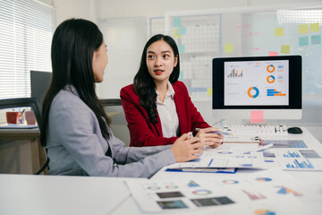 Two asian businesswomen discussing financial charts and data on a computer screen, analyzing company performance and making strategic decisions during an office meeting