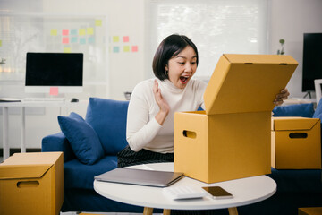 Young asian woman is sitting on a blue sofa and opening a cardboard box. She is surprised by the...
