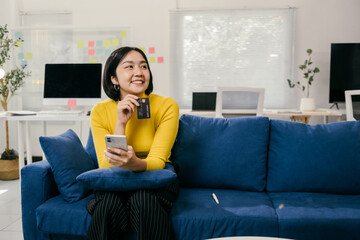 Young asian woman is holding credit card and using smartphone for online shopping on sofa at home,...