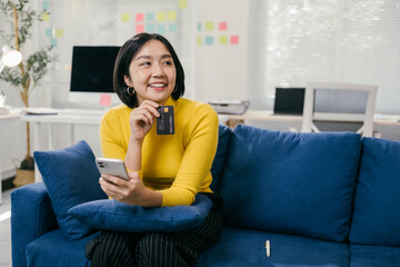 Young woman happily shops online at home, holding a credit card and using her phone, enjoying the...