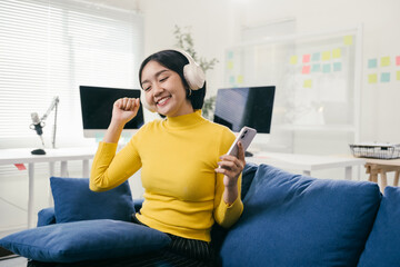 Young asian woman is sitting on a blue couch listening to music on her headphones and holding her smartphone. She is smiling and dancing