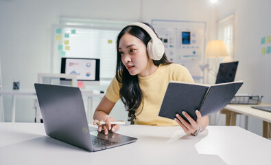 Young asian woman wearing headphones is using a laptop and taking notes while participating in an online course at her home office