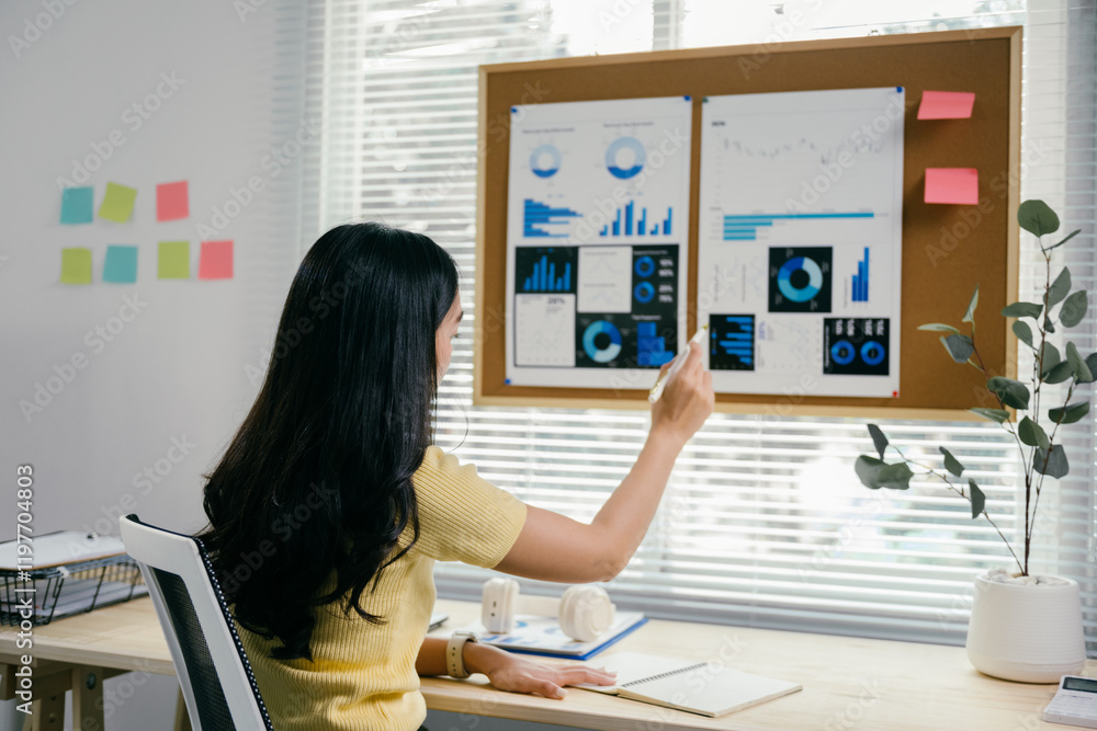 Poster young asian businesswoman is pointing at a chart on a board and analyzing data while sitting at her 