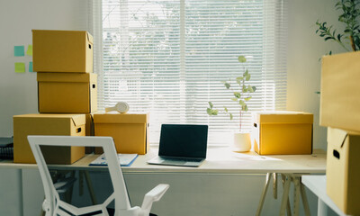 Cozy home office with neatly stacked cardboard boxes on a desk, bathed in sunlight. Laptop, chair, and houseplant add a personal touch, symbolizing new beginnings and change
