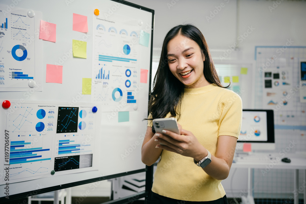 Poster businesswoman is smiling while looking at her smartphone in an office with charts and graphs on a wh