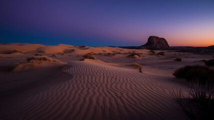 Naklejka premium Vast Desert Landscape at Twilight with Endless Golden Dunes, Sharp Shadows, and a Solitary Rock Formation Under a Deep Purple to Orange Sky