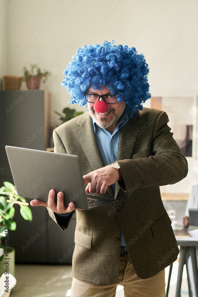 Poster happy mature boss in clown wig looking at camera in office while using laptop and pointing at screen