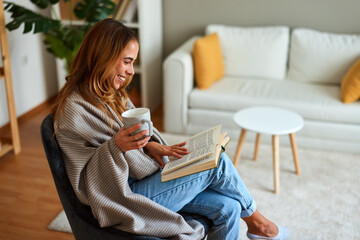 Young woman enjoying a cup of tea while reading a book sitting comfortably on an armchair at home...