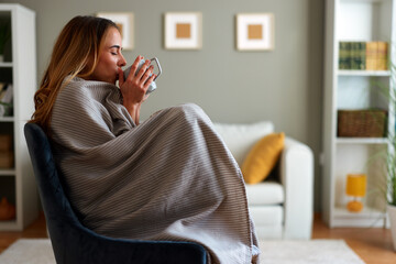 Woman sitting on armchair wrapped in a blanket enjoying a hot beverage in the comfort of her home