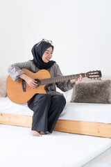 happy and playful asian moslem woman plays guitar in her bedroom. muslim girl play music instrument while smiling