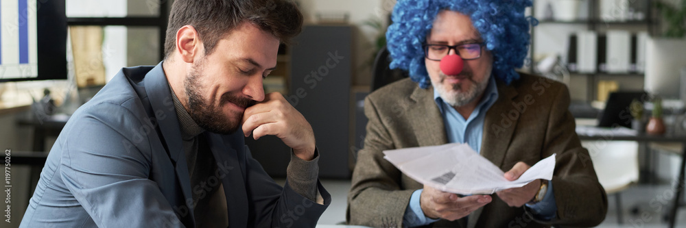 Aufkleber header of young smiling male employee sitting by workplace next to clown boss and looking through fi