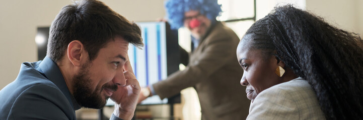 Header of young smiling subordinates looking at one another while making fun of their boss wearing...