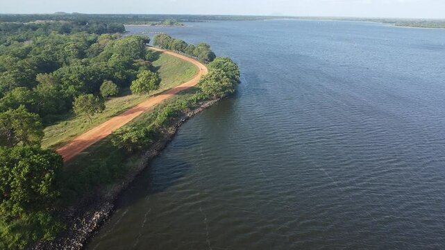 Aerial footage of a gravel road and green fields adjacent the Mahakanadara Wewa reservoir, Sri Lanka