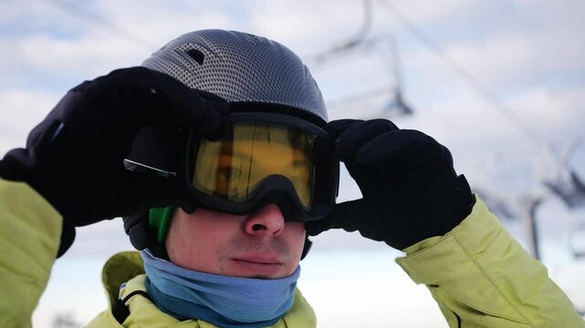 Happy male skier puts on ski goggles and helmet, preparing for a day on the slopes at a ski resort, with ski lifts visible in the background