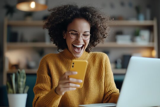 A cheerful young woman is using her smartphone while sitting at a desk with a laptop