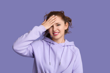 Portrait of ashamed young woman covering face with hand on purple background