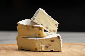 Wheel of creamy camembert cheese topped with nuts, resting on a rustic wooden surface against a solid black background. The rich textures and contrast create an elegant culinary composition